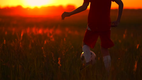 Close-up-of-foot-boy-football-player-running-at-sunset-with-a-ball-in-the-field-camera-moves-behind-the-boy.